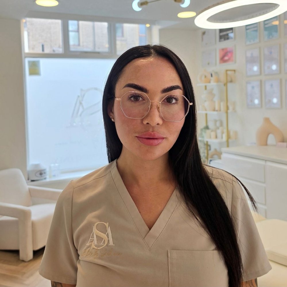 Sarah Ann Roberts standing in her Oldham clinic in ASA branded scrubs, with framed certificates on the wall behind her.